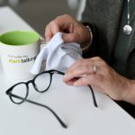 Detailaufnahme einer Person am Schreibtisch, die eine Brille mit einem Tuch reinigt. Daneben steht eine Kaffeetasse mit der Aufschrift „start:talking“. Das Bild steht für ruhiges Arbeiten, Fokus und Klarheit in der digitalen B2B-Kommunikation.