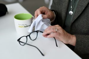 Detailaufnahme einer Person am Schreibtisch, die eine Brille mit einem Tuch reinigt. Daneben steht eine Kaffeetasse mit der Aufschrift „start:talking“. Das Bild steht für ruhiges Arbeiten, Fokus und Klarheit in der digitalen B2B-Kommunikation.